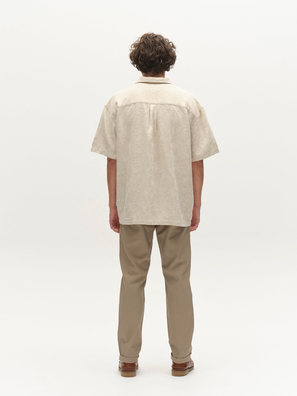 A person with curly hair stands facing away against a plain white background, wearing a short-sleeved beige shirt, brown shoes, and Gabba's GABpaul L.C. Zip Crop Doby Pant in Sand.