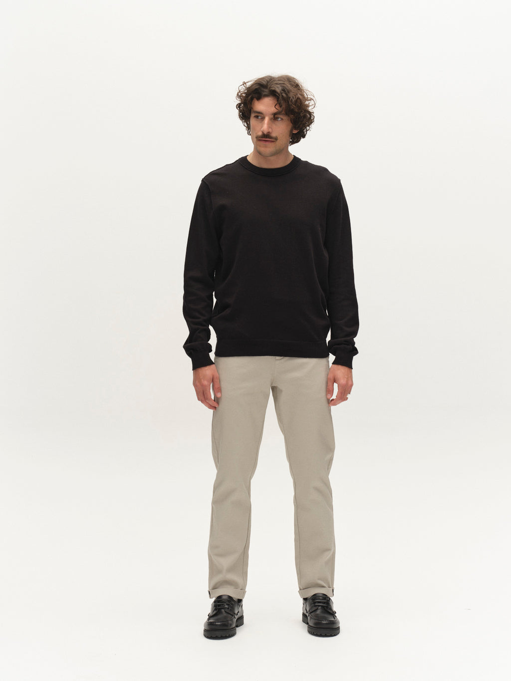 A man with curly hair and a mustache stands facing forward in a black long-sleeve shirt, Gabba GABpaul L.C. Zip Crop Doby Pant in Lt. Sand, and black shoes against a plain white background.