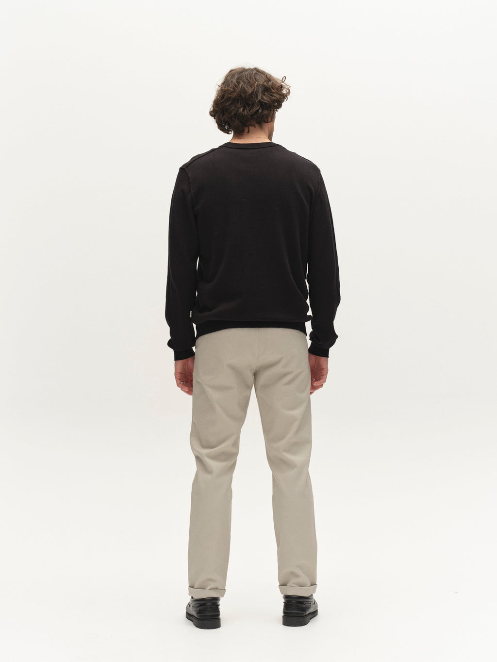 A person with curly hair stands against a white background, wearing a black long-sleeve shirt, black shoes, and Gabba’s GABpaul L.C. Zip Crop Doby Pant in Lt. Sand.