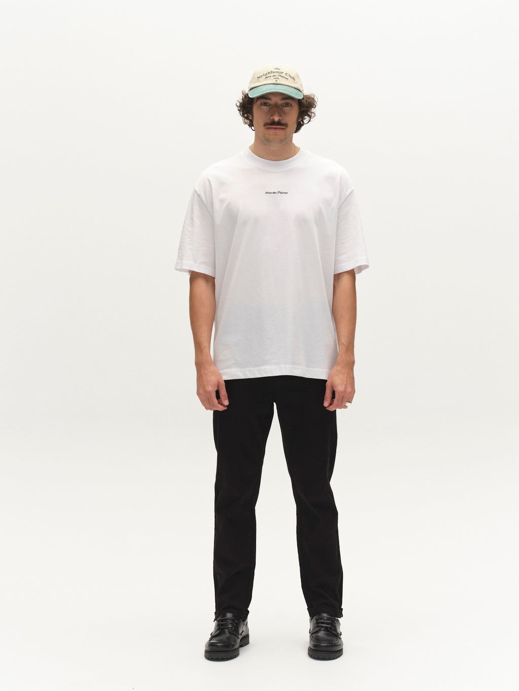 A man with curly hair and a mustache stands facing forward against a white background, wearing a beige baseball cap, loose white T-shirt, black shoes, and Gabba GABpaul L.C. Zip Crop Doby Pant in Blue Black.