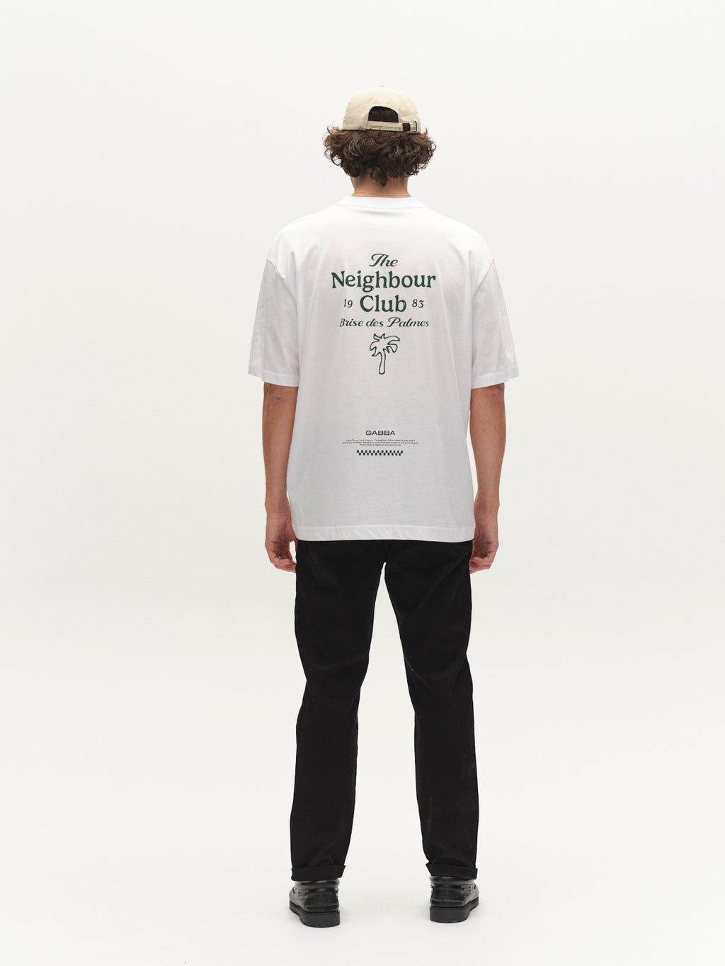 A person, facing away in a light studio, wears a white cap, white T-shirt with back text, and Gabba GABpaul L.C. Zip Crop Doby Pant in Blue Black with black shoes.