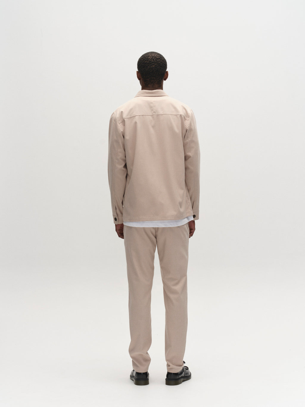 A person stands facing away from the camera against a plain white background, wearing the Gabba GABtogger Base Deme LS Overshirt in Birch with matching pants and a white shirt—an effortless choice for seasonal layering.