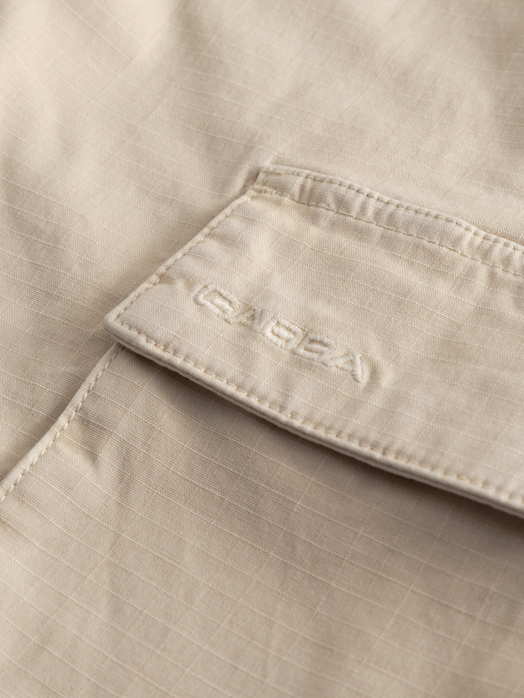 Close-up of the GABjoli Cargo Ribstop Shorts - Birch by Gabba, showing a beige fabric pocket with subtle grid patterns and GARBA embroidery on the flap, highlighting the relaxed-fit cargo pockets for versatile everyday wear.