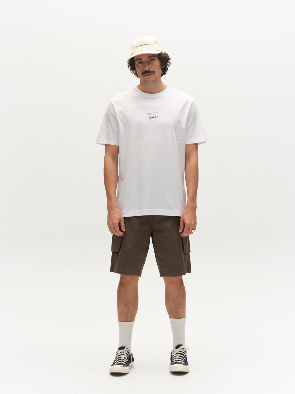 A man with a mustache stands against a plain white background, wearing a white T-shirt and Gabba's GABjoli Cargo Ripstop Shorts in Army—relaxed-fit shorts with side cargo pockets—paired with white socks, black sneakers, and a beige cap.