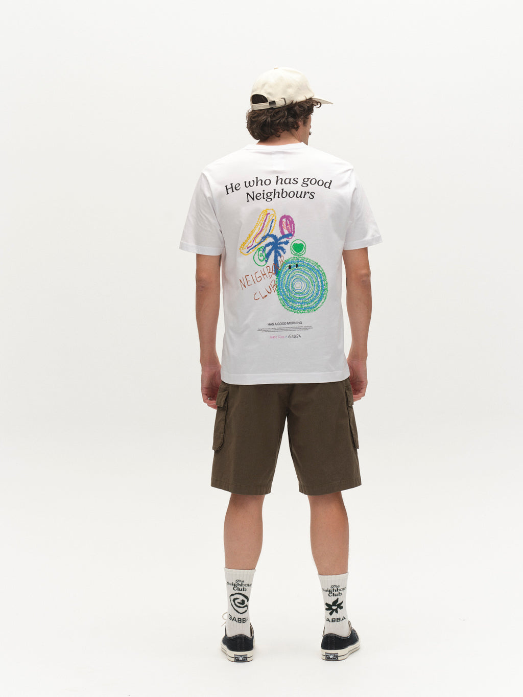 A person faces away, wearing a white graphic T-shirt, Gabba GABjoli Cargo Ripstop Shorts in Army with side cargo pockets, white socks with black designs, sneakers, and a light-colored cap. The background is white.