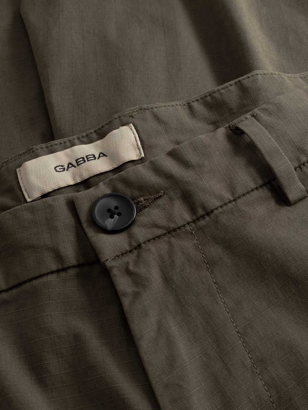 Close-up of Gabba GABjoli Cargo Ripstop Shorts in army green, featuring a black button, belt loop, and a white GABBA label inside the waistband. The subtle grid-textured fabric enhances this versatile everyday look.