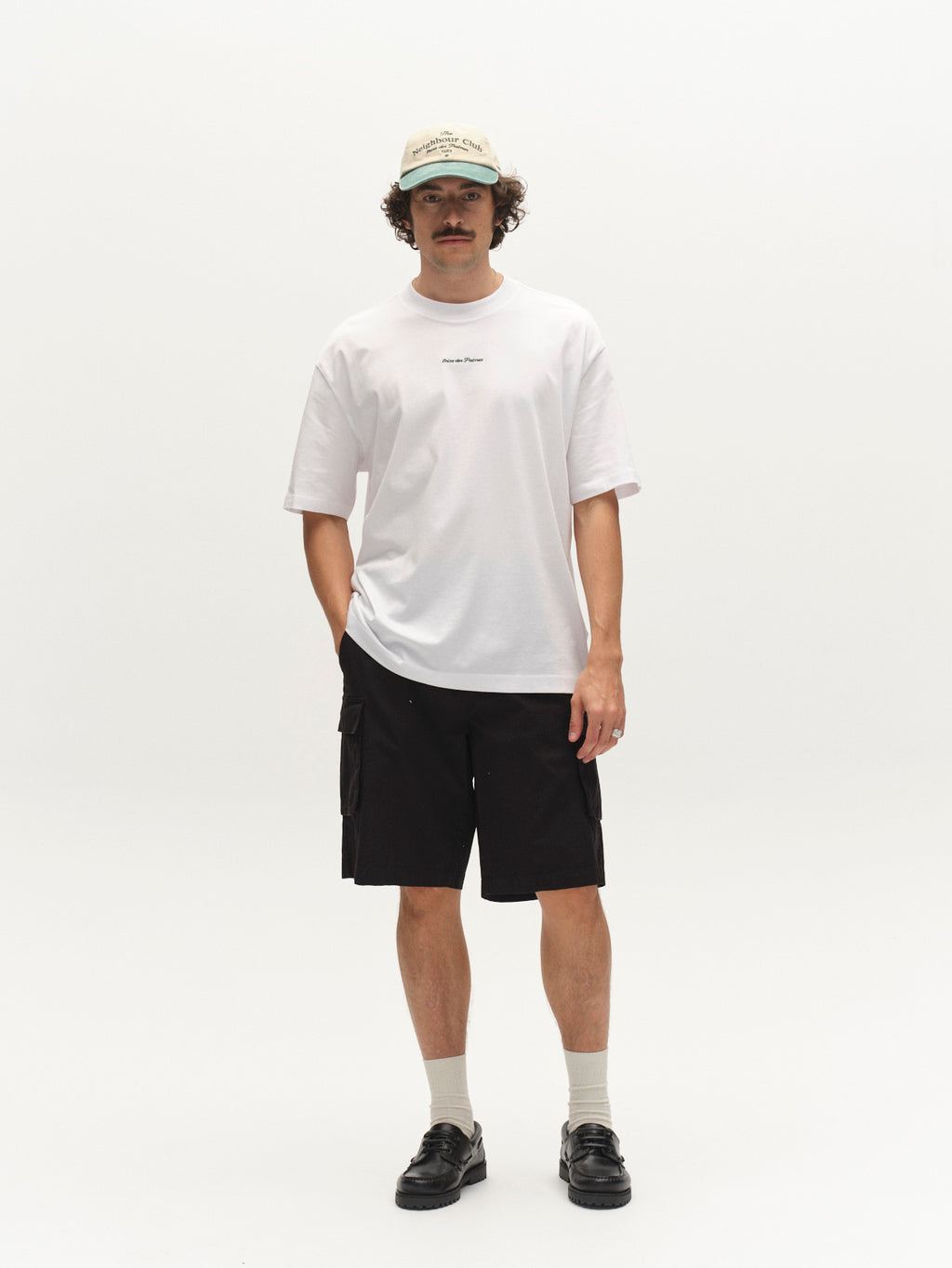 A man with a mustache and curly hair stands against a white background, wearing a beige cap, white t-shirt, and Gabba GABjoli Cargo Ribstop Shorts in black—relaxed fit with side cargo pockets for versatile everyday style.