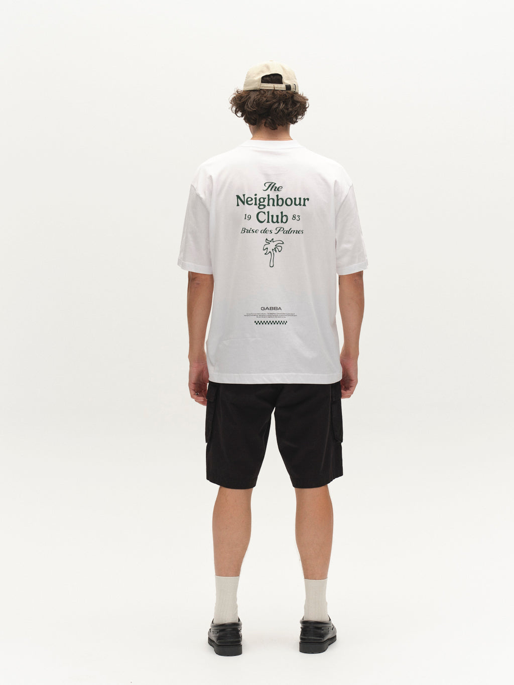 A person with curly hair, wearing a beige cap, white printed tee, Gabba GABjoli Cargo Ribstop Shorts - Black, white socks, and black shoes stands facing away in a plain white studio for versatile everyday style.