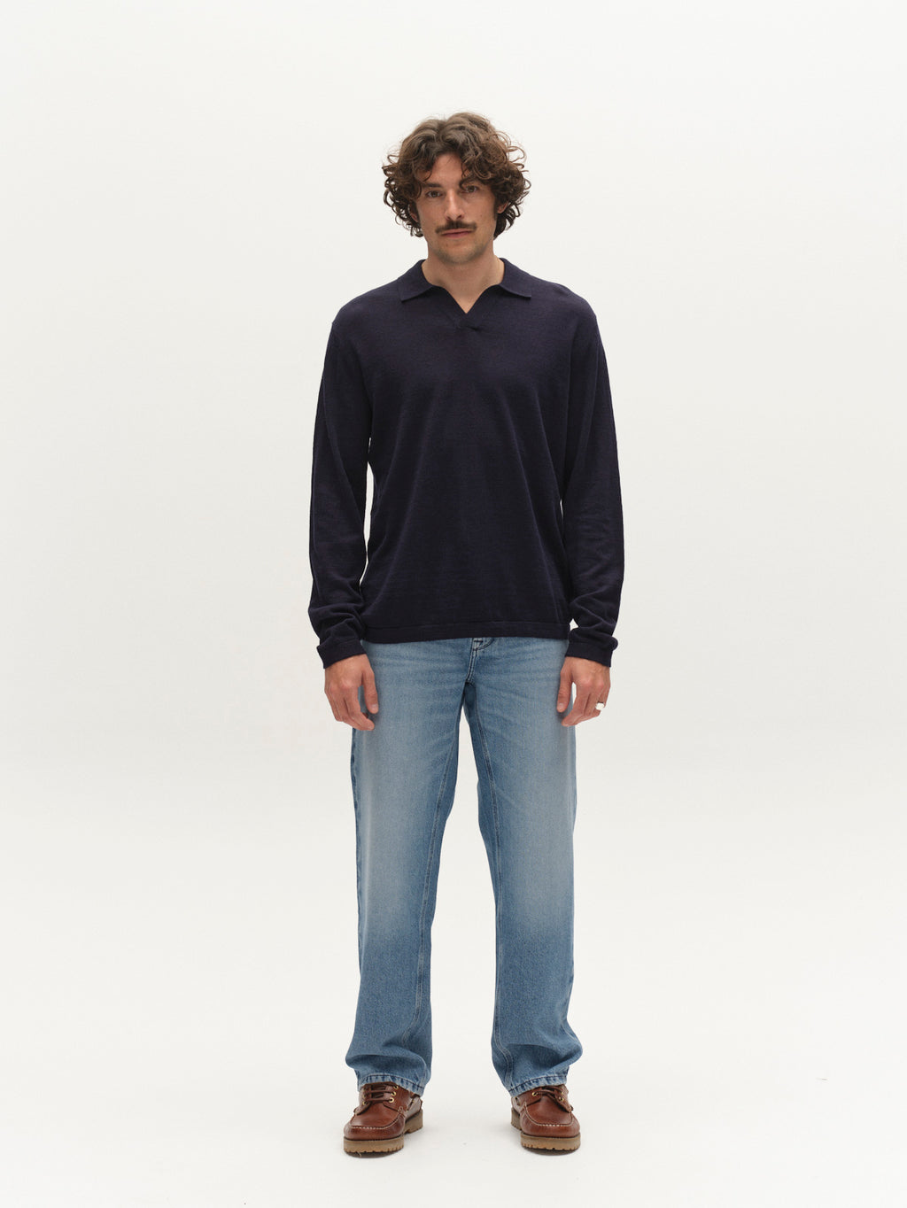 A man with curly hair and a mustache stands against a plain white background, wearing the Gabba GABcross Dush LS Knit in navy, paired with light blue jeans and brown shoes.