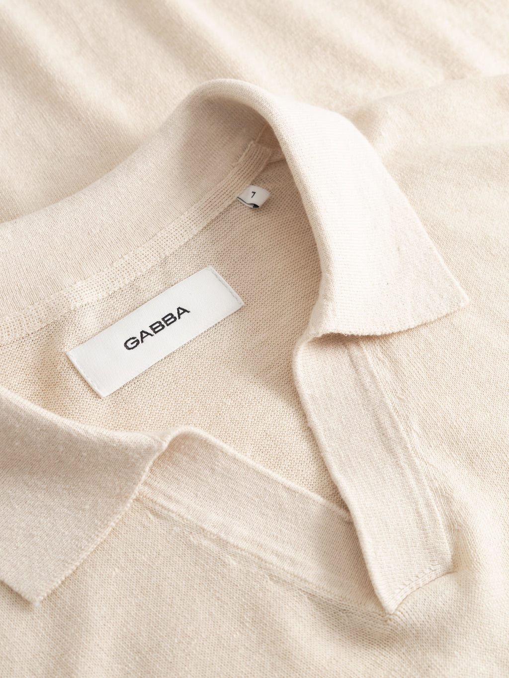 Close-up of the GABcross Dush LS Knit in Sand Mel., a beige Gabba shirt made from soft fabric with a relaxed collar and an inner label showing the Gabba brand and size L.