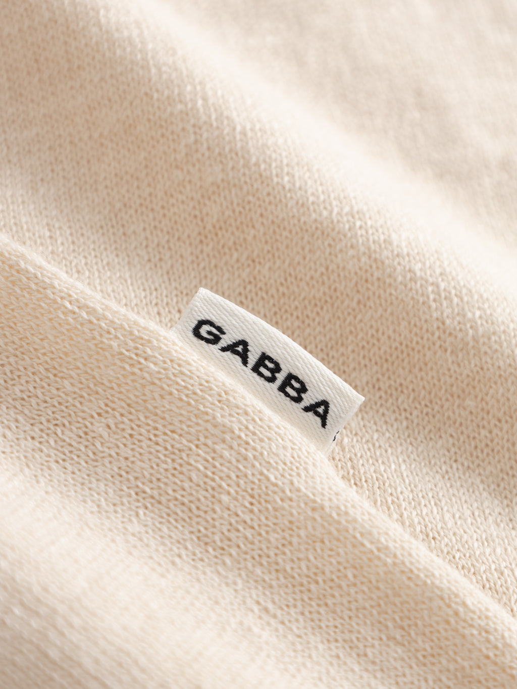 A close-up of the GABcross Dush LS Knit in Sand Mel by Gabba, featuring beige flat knit fabric and a small white tag with 