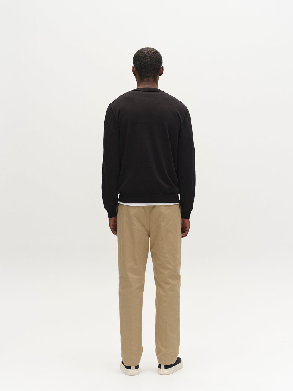 A man stands facing away from the camera against a plain white background, wearing the Gabba GABfumo Spy LS Knit sweater in black, beige pants, and black-and-white shoes.