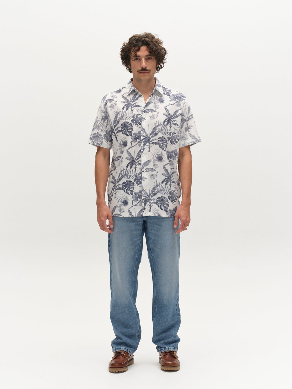 A man with curly hair and a mustache faces forward, wearing the Gabba GABsven Reg Leaves SS Shirt - Flower Mix, paired with light blue jeans and brown shoes, standing against a plain white background.