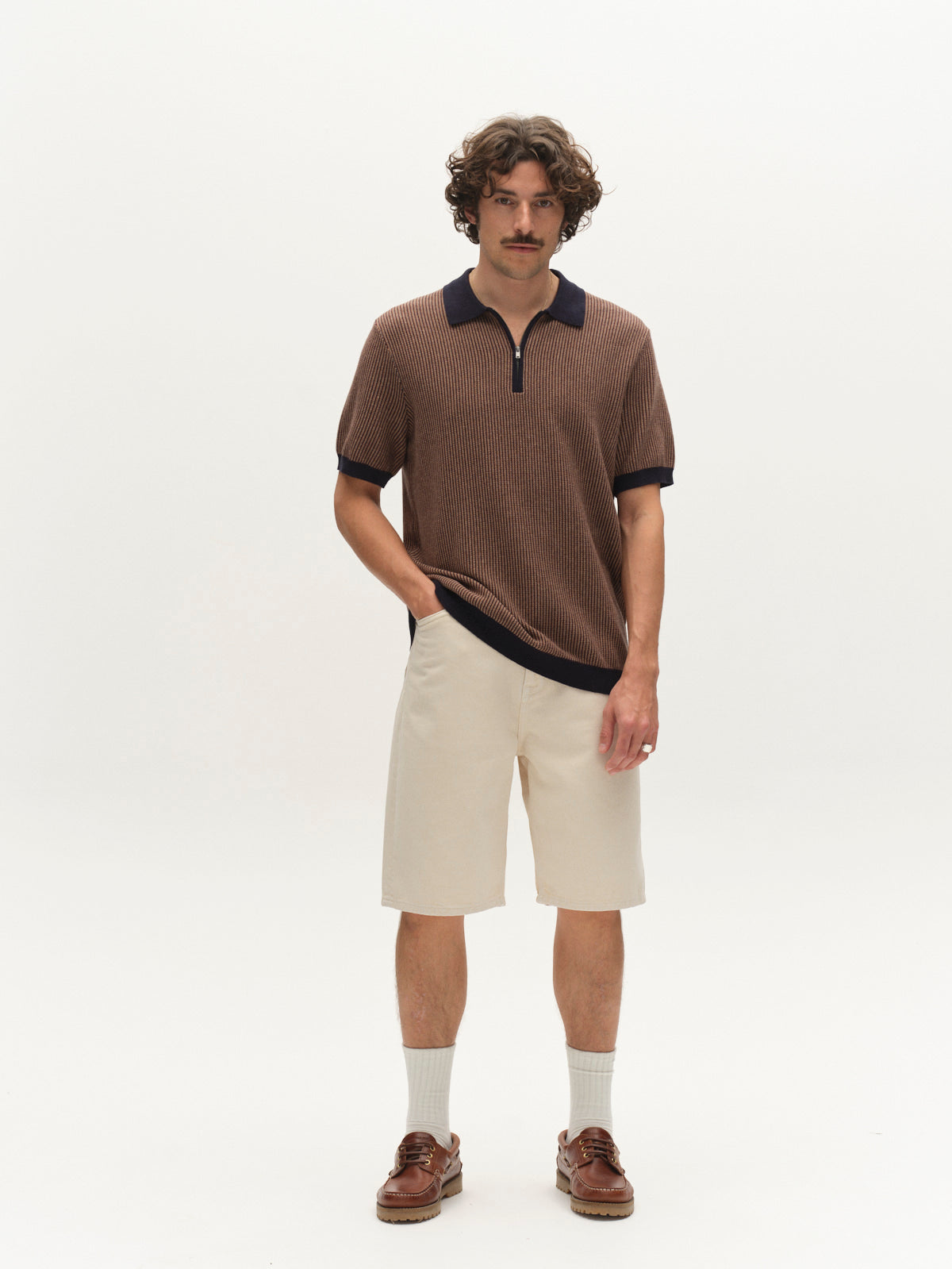 A man with curly hair and a mustache wears a brown knit polo, Gabba GABmox Antiq Shorts in Ecru, light socks, and brown loafers while standing against a plain white background.