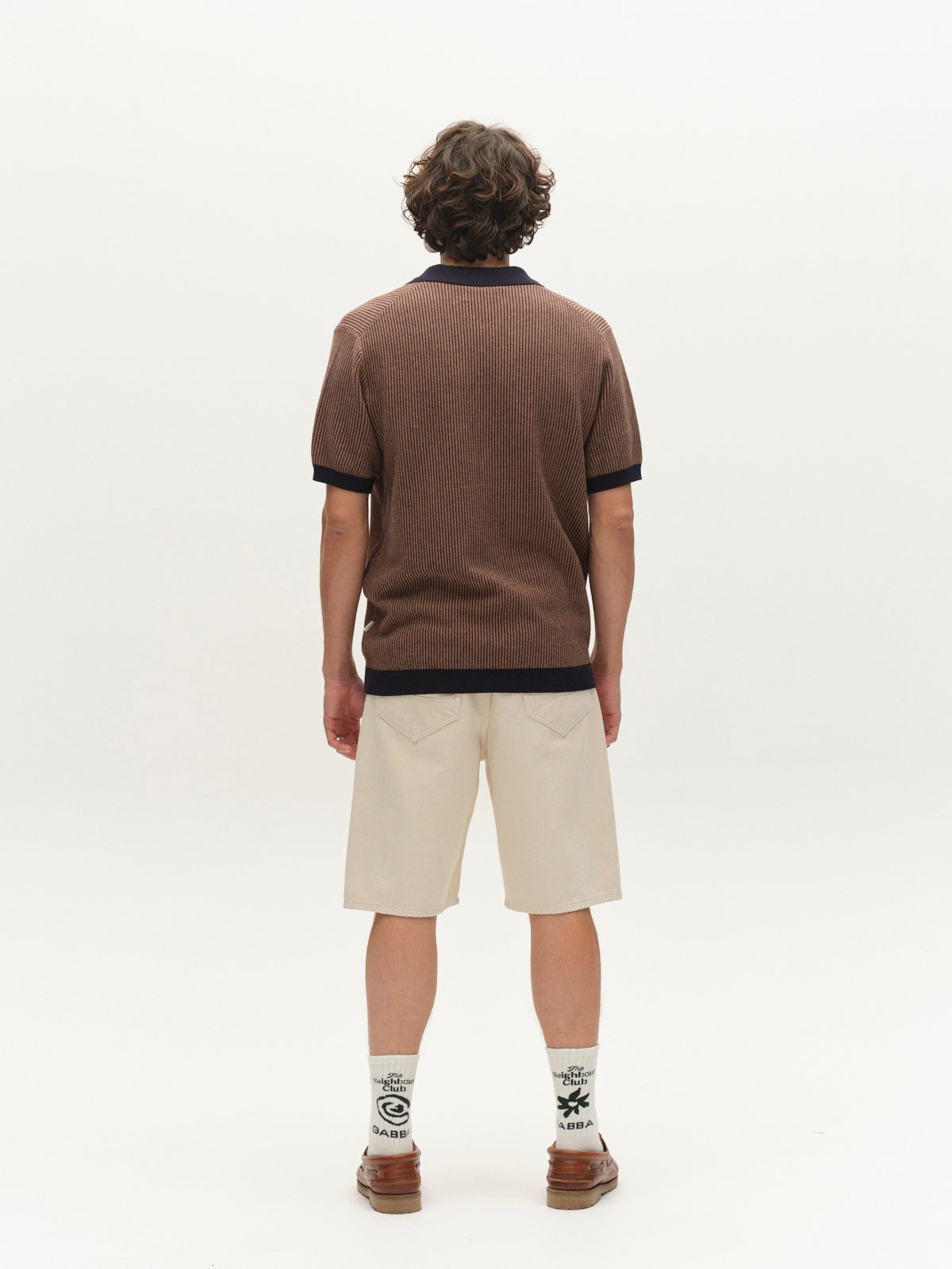 A person with curly hair stands facing away, wearing a brown short-sleeve collared shirt, Gabba GABmox Antiq Shorts in Ecru, printed white socks, and brown shoes, set against a plain white background.