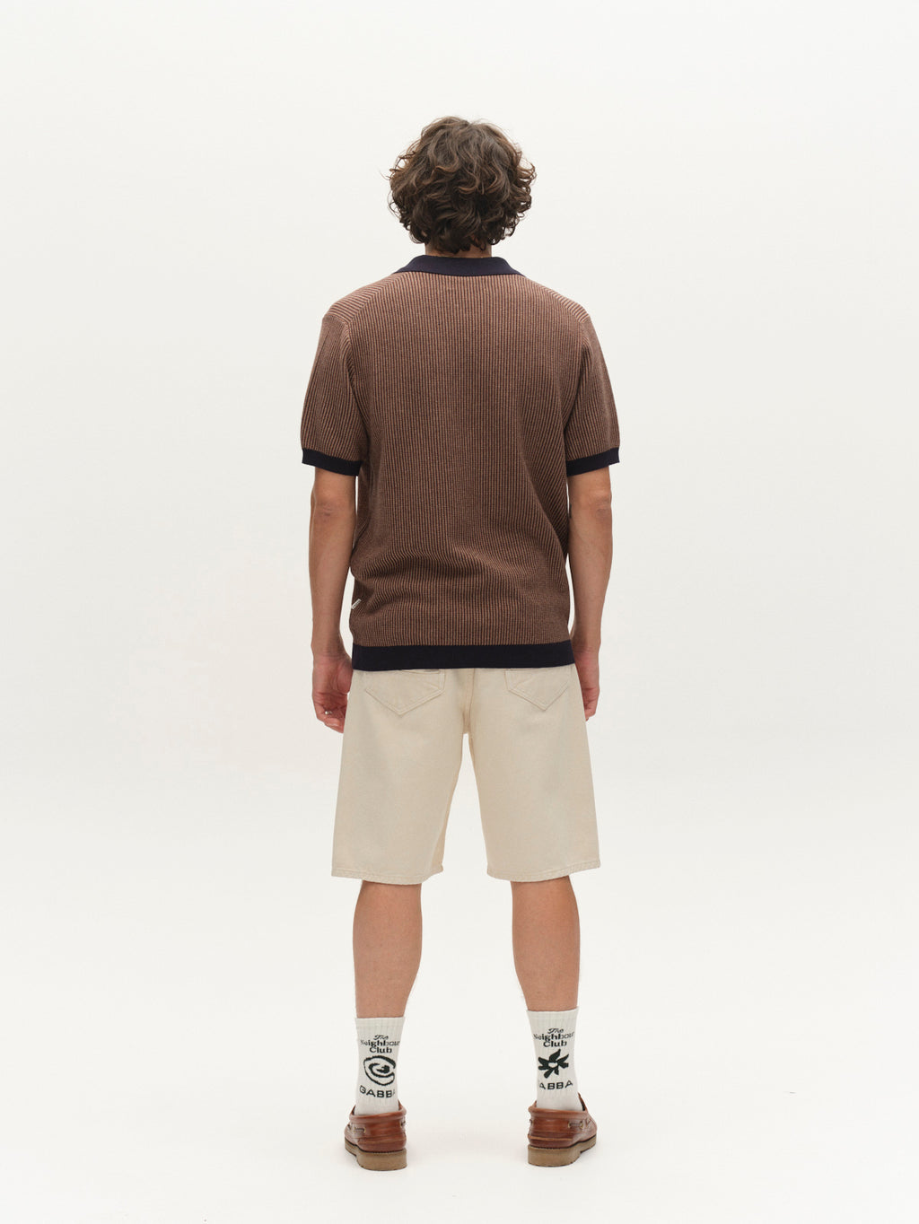A person with curly hair stands facing away, wearing a brown short-sleeve collared shirt, Gabba GABmox Antiq Shorts in Ecru, printed white socks, and brown shoes, set against a plain white background.