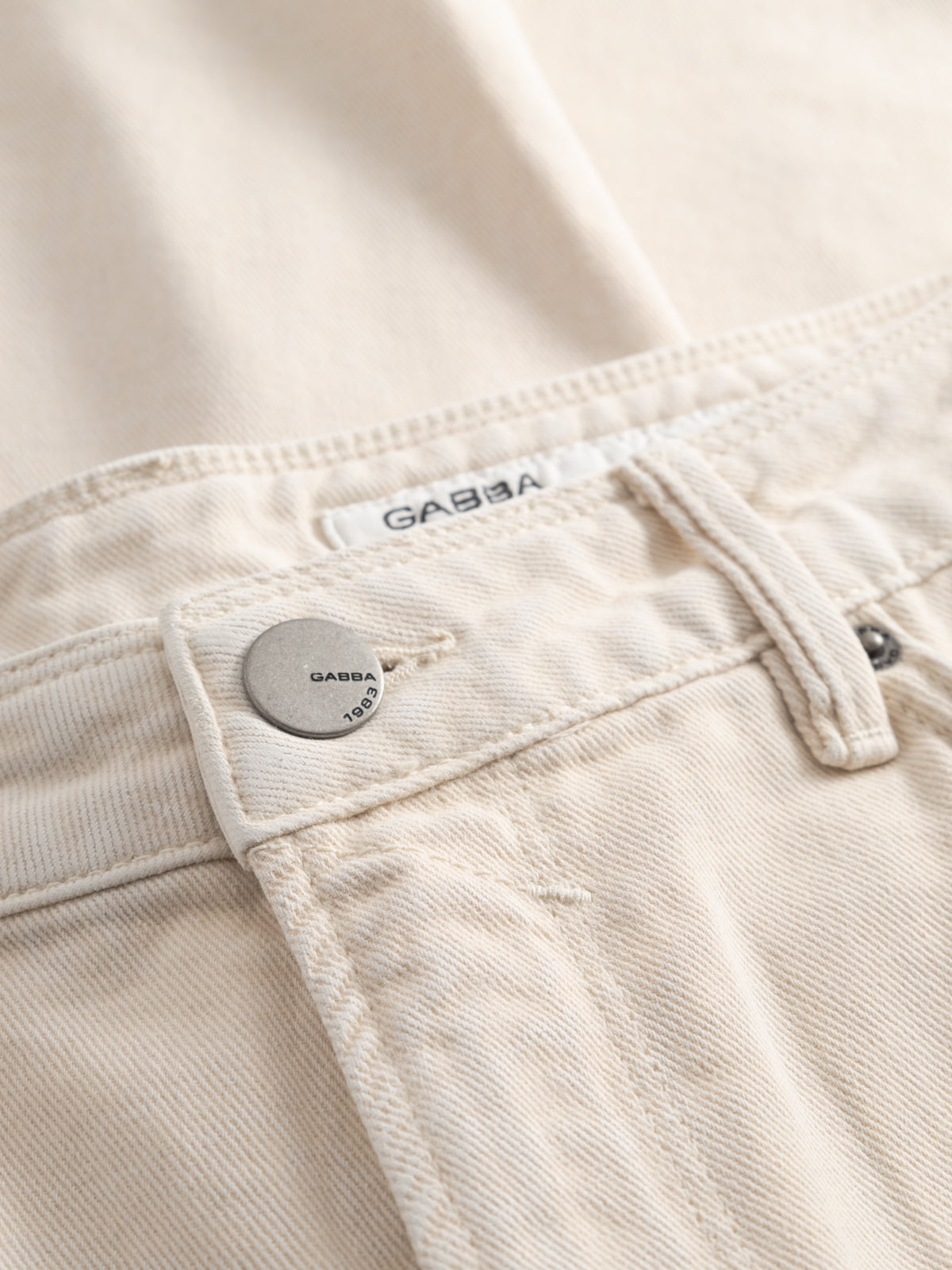 Close-up of GABmox Antiq Shorts in ecru denim by Gabba, featuring rigid twill fabric, a waistband with a metal button engraved 