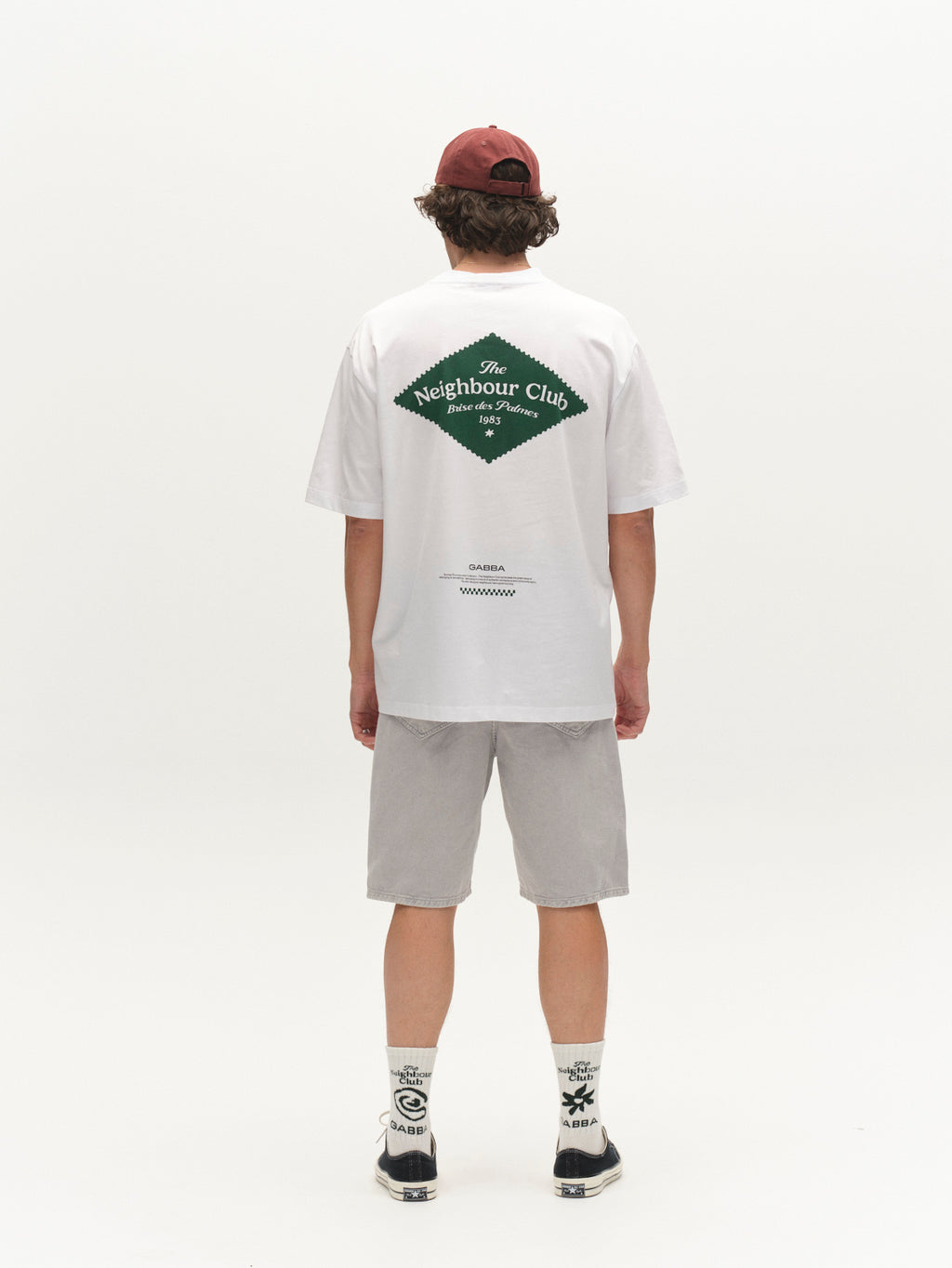 A person stands facing away, wearing a maroon cap, white tee with The Neighbour Club logo on the back, Gabba GABmox Antiq Shorts in Denim Blue, white socks with black accents, and beige sneakers against a plain white background.