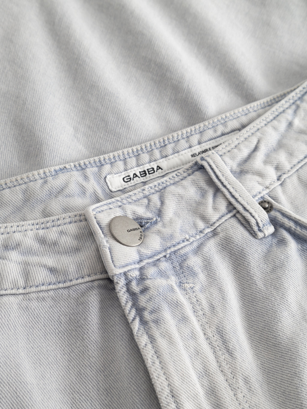 Close-up of GABmox Antiq Shorts in denim blue by Gabba, featuring a visible Gabba label on the waistband and a metallic brand-engraved button. Crafted from rigid twill, the shorts are laid flat on matching denim.