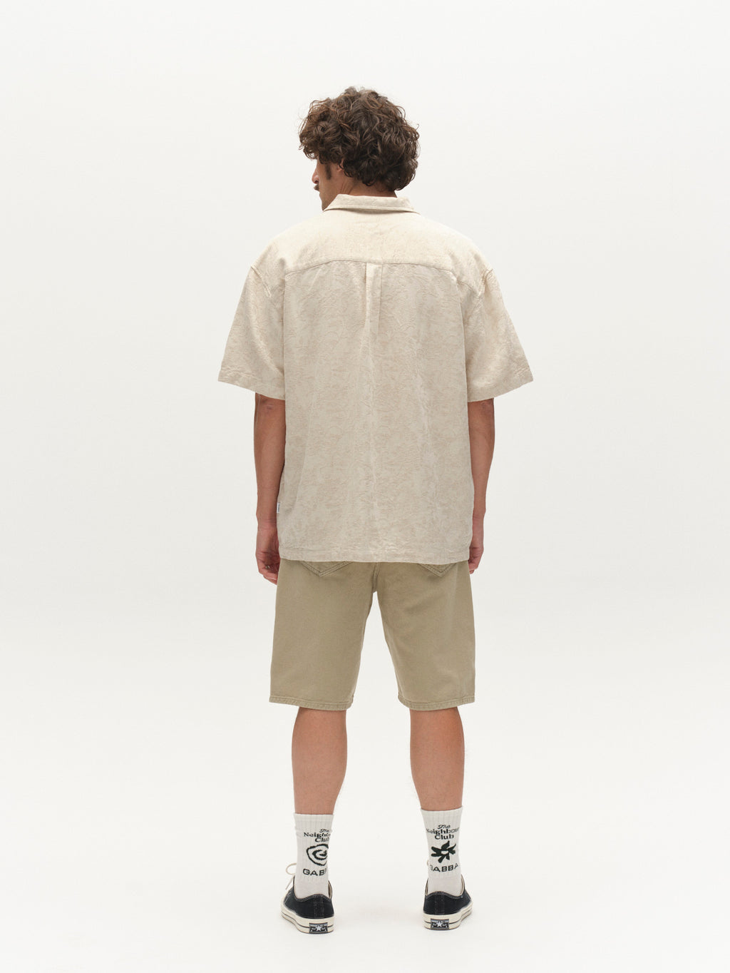 Facing away, a person with curly hair wears a light beige short-sleeve shirt and Gabba GABmox Antiq Shorts in Alfa Green, paired with white socks featuring black designs and black sneakers, against a plain white background.