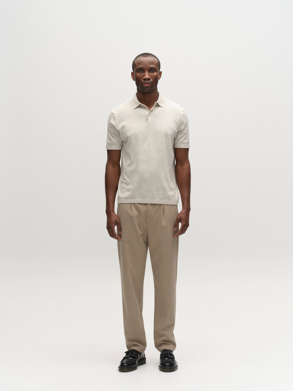 A man faces the camera against a plain white background, wearing the Gabba GABbenson New Cot SS Knit - Sand Mel polo shirt, tan trousers, and black shoes.