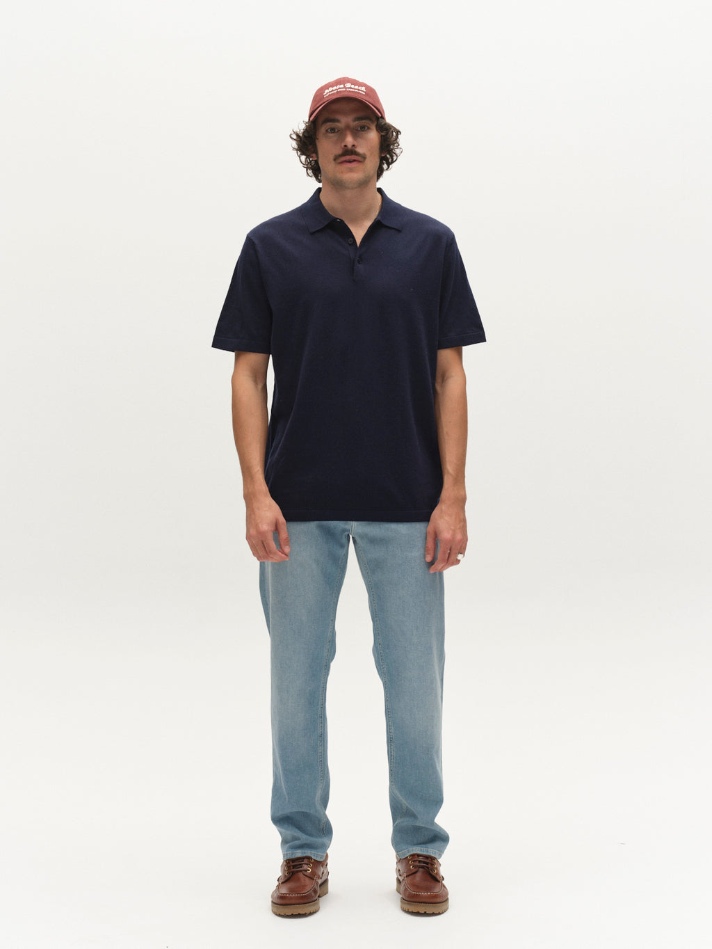 A man with a mustache faces forward against a plain white background, wearing a maroon cap, Gabba GABbenson New Cot SS Knit polo in navy melange, light blue jeans, and brown shoes.