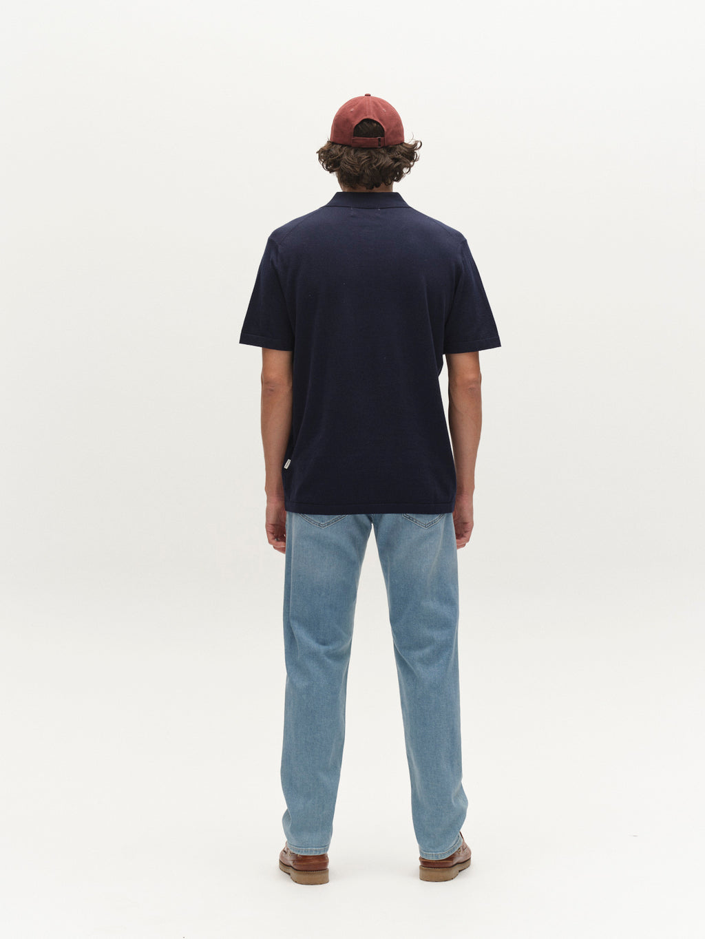 A person stands with their back to the camera, wearing a maroon cap, light blue jeans, brown shoes, and a Gabba GABbenson New Cot SS Knit short-sleeve polo in Navy Mel. The background is plain white.