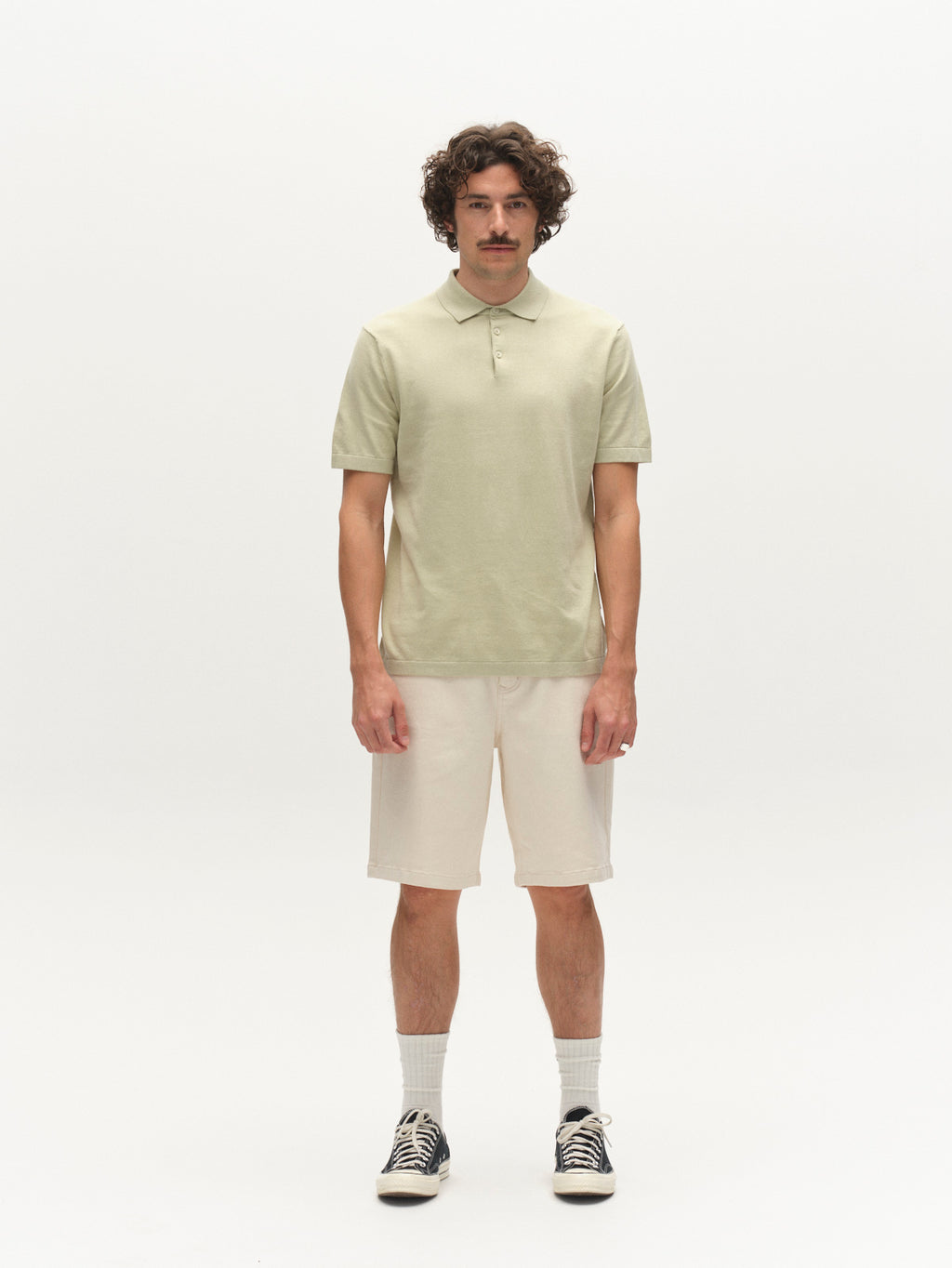 A man with curly hair and a mustache faces forward, wearing the Gabba GABbenson New Cot SS Knit polo in Alfa Green, beige shorts, gray socks, and black sneakers. The background is plain white.