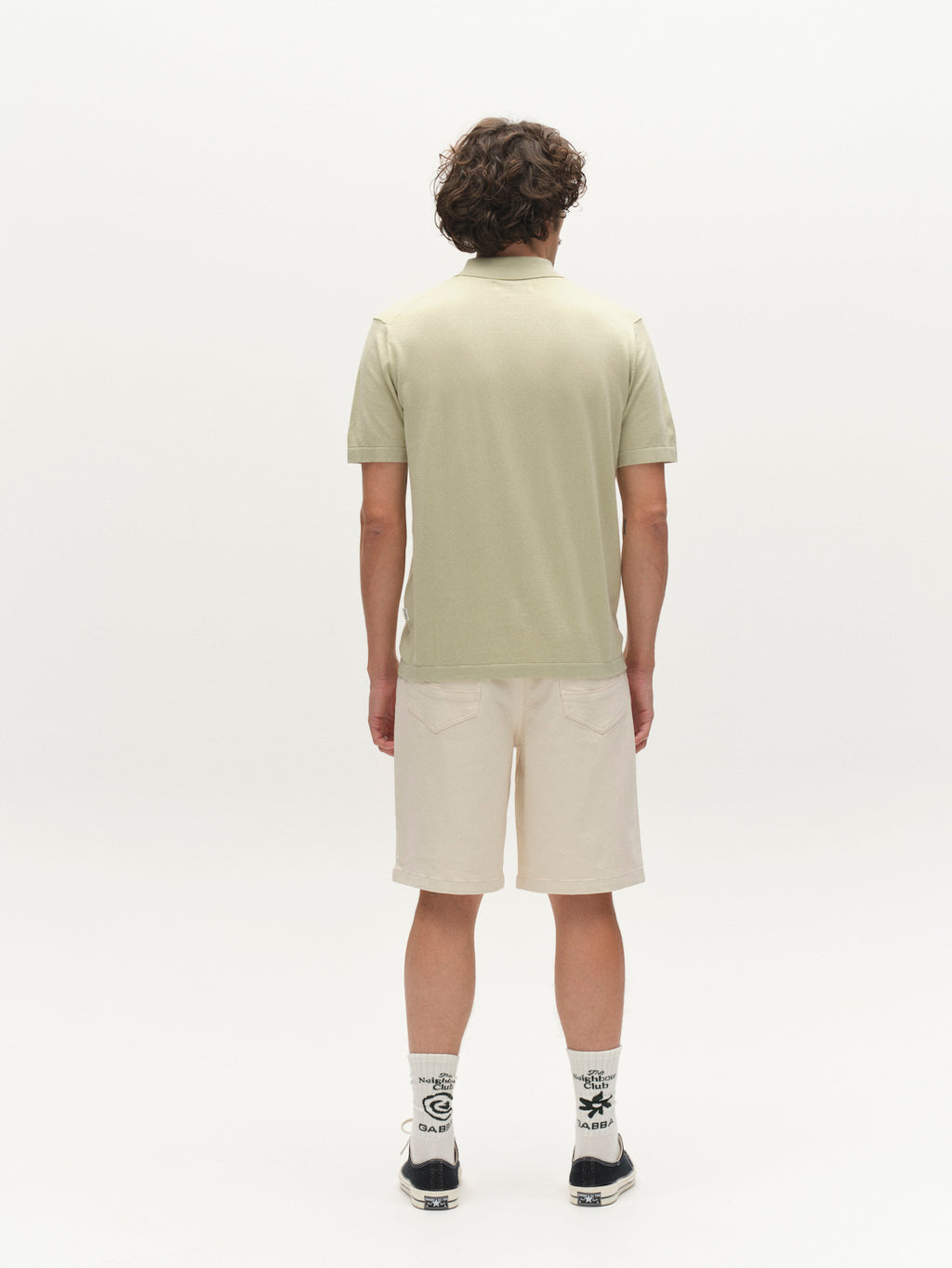 A person with curly brown hair, facing away from the camera, wears a Gabba GABbenson New Cot SS Knit in Alfa Green, beige knee-length shorts, white socks with black designs, and black sneakers against a plain white background.