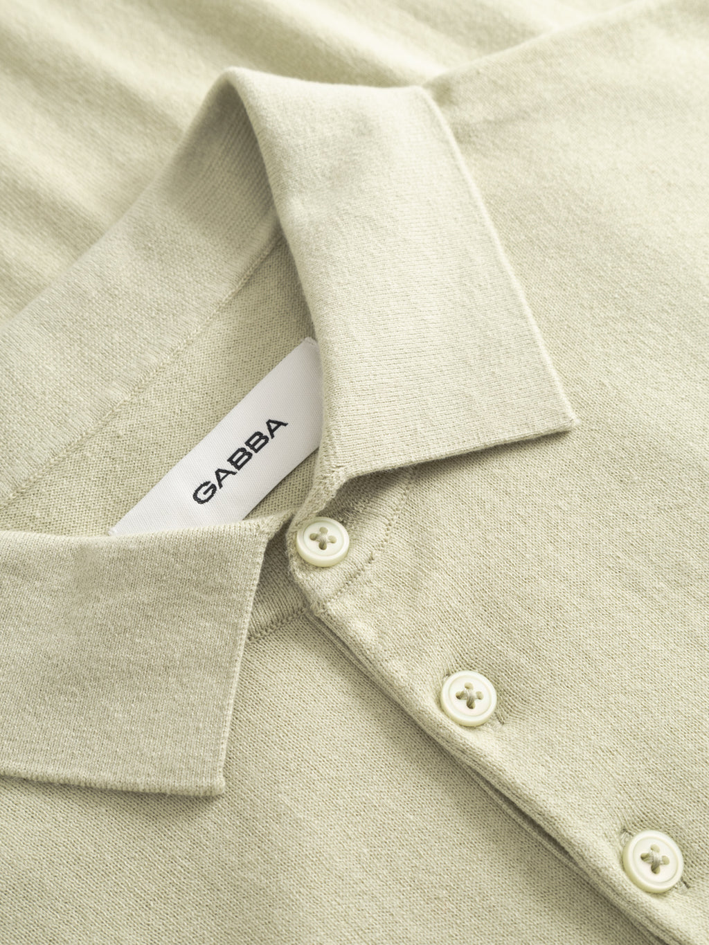 Close-up of the GABbenson New Cot SS Knit in Alfa Green by Gabba, featuring a folded collar, three white buttons, and an inner neckline label that reads Gabba.