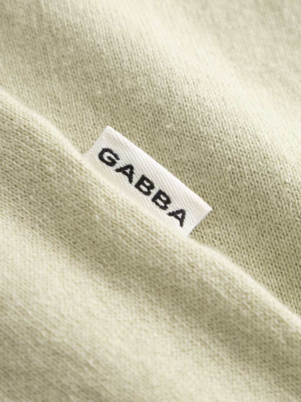 A close-up of the soft, Alfa Green GABbenson New Cot SS Knit fabric by Gabba shows a white tag, partially tucked under a fold, with 