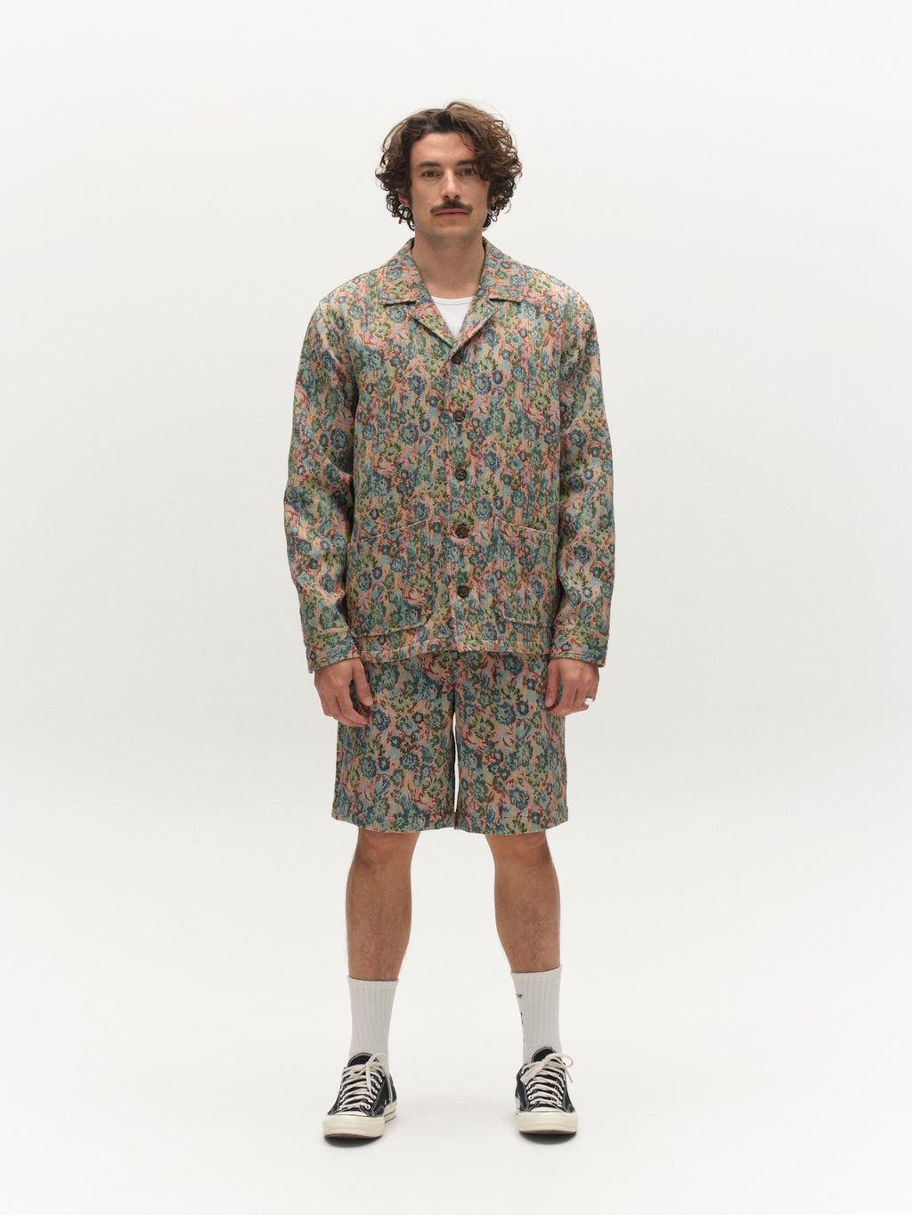 A man with curly hair and a mustache stands against a white background, sporting Gabba’s GABjoli Ted Kscope Shorts - Abstract Flowers with a matching jacket, white crew socks, and black sneakers for effortless seasonal style.