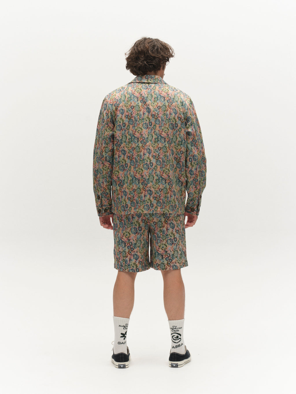 A person stands facing away, wearing Gabba's GABjoli Ted Kscope Shorts - Abstract Flowers and a matching floral long-sleeve shirt, paired with white socks featuring black text and black shoes, against a plain white background.
