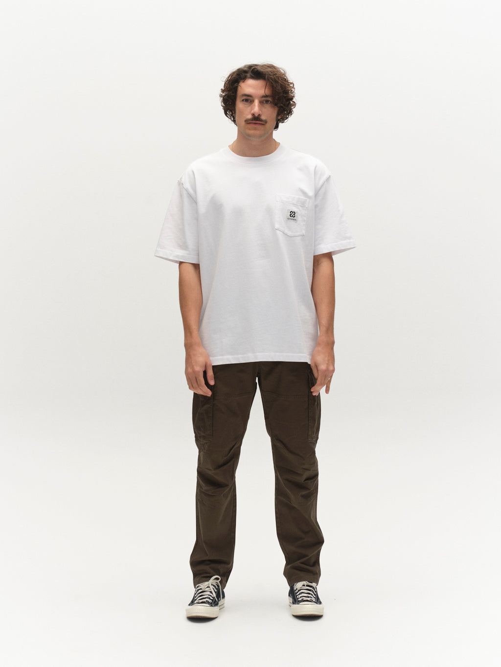 A man with curly hair and a mustache stands facing forward against a plain white background, wearing a loose white t-shirt, Gabba GABjoe Vico Cargo Pant - Army, and black sneakers.