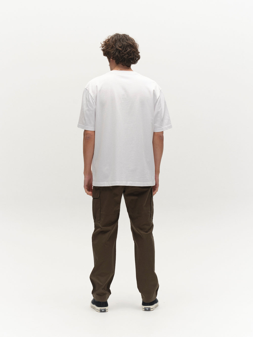 A person with curly hair stands facing away, wearing a plain white t-shirt, black shoes, and Gabba GABjoe Vico Cargo Pant in Army green against a plain white background.