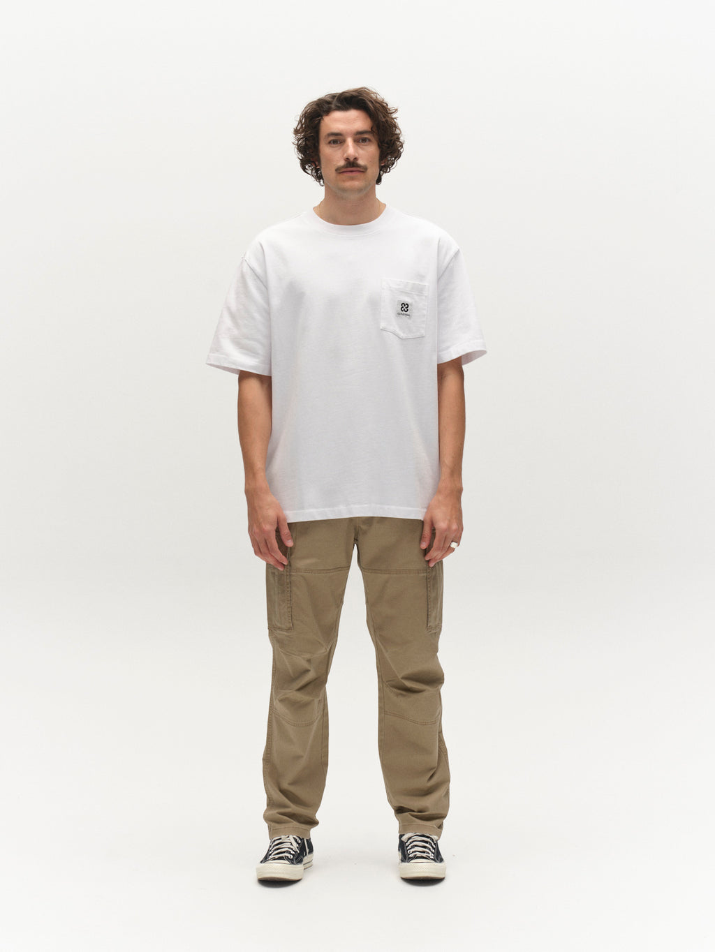A man with wavy hair and a mustache faces forward in a white tee with chest pocket, black-and-white sneakers, and Gabba GABjoe Vico Cargo Pant in Sand, standing against a plain white background.