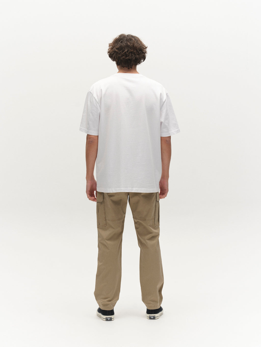 A person with short, curly hair faces away from the camera in a plain white t-shirt and Gabba GABjoe Vico Cargo Pant in Sand, paired with black sneakers, against a plain white background.