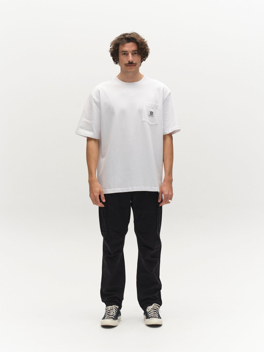 A man with curly hair and a mustache faces forward, wearing a loose white T-shirt with a pocket, Gabba GABjoe Vico Cargo Pant in black, and striped sneakers against a plain white background.