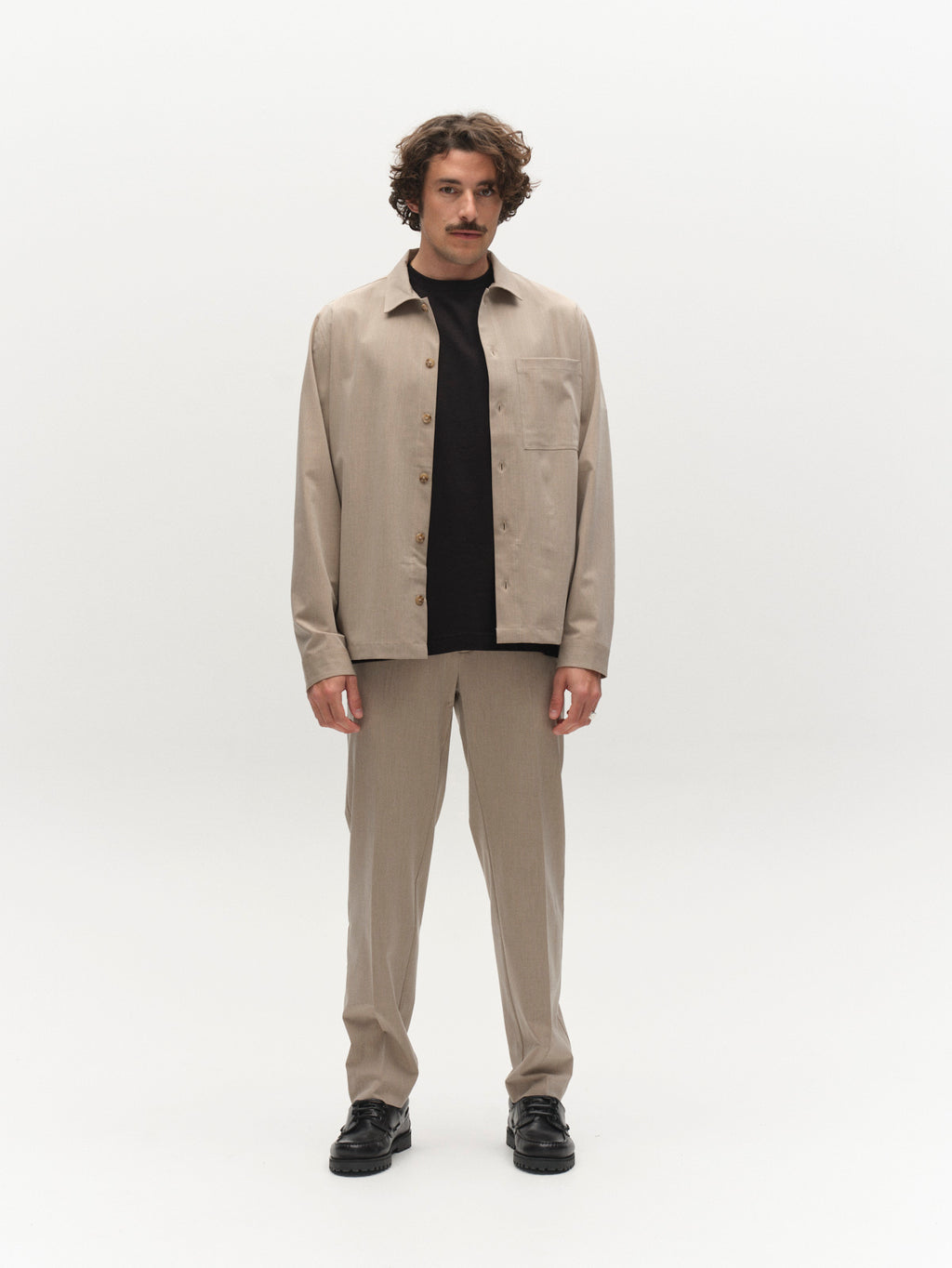 A man with wavy hair and a mustache stands against a white background wearing the Gabba GABtogger Tailor Dressy Overshirt in sand over a black t-shirt, paired with beige trousers and black shoes for smart layering.