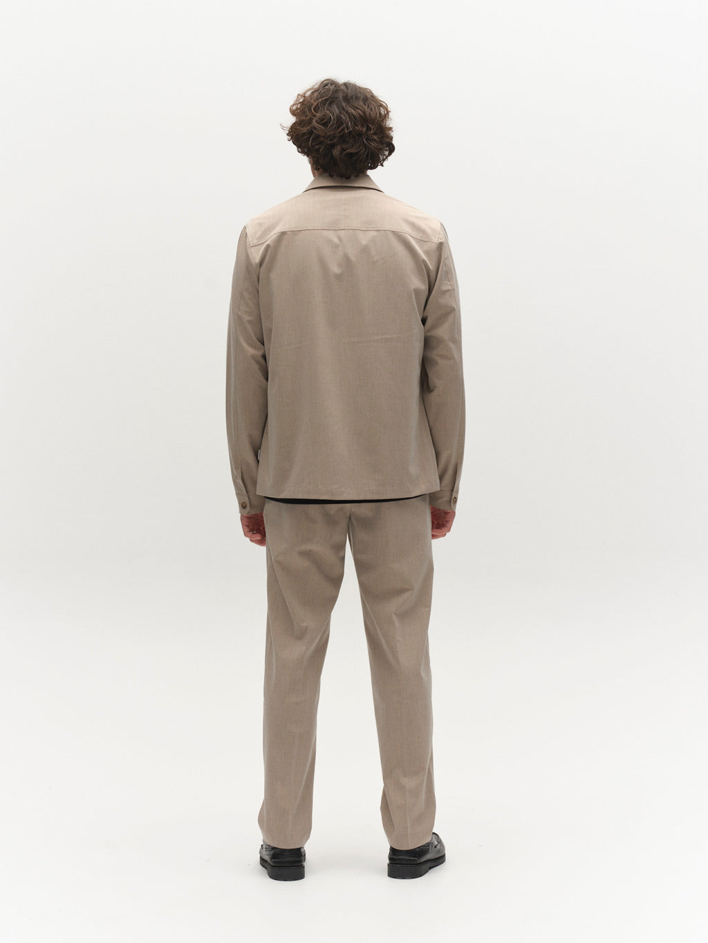 A person with curly brown hair stands facing away from the camera, layering the Gabba GABtogger Tailor Dressy Overshirt in Sand, paired with black shoes, against a plain white background.
