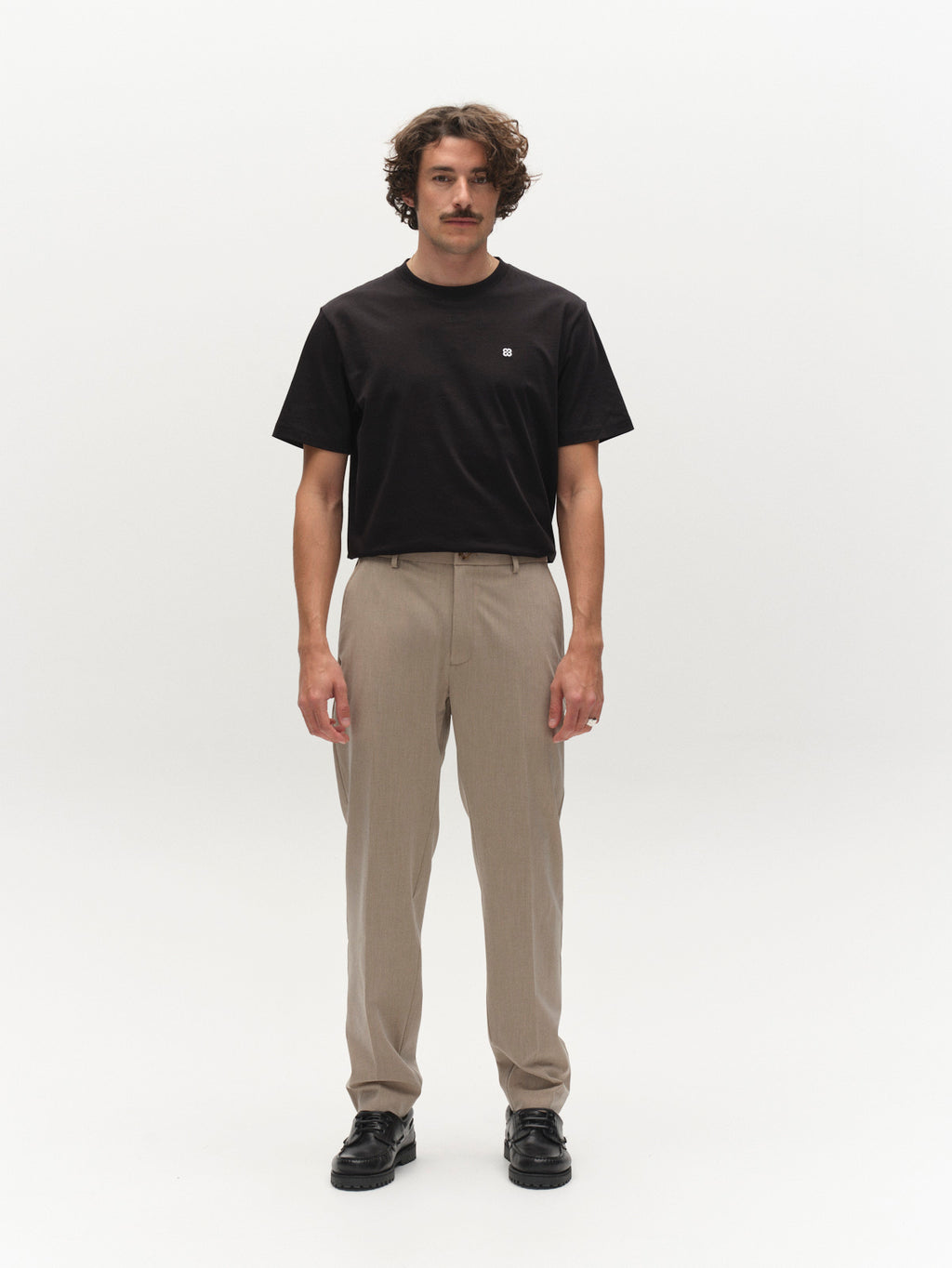 A man with curly hair and a mustache stands facing forward against a plain white background, wearing a black t-shirt, black shoes, and Gabba GABjoe Tailor Dressy Pant in Sand.