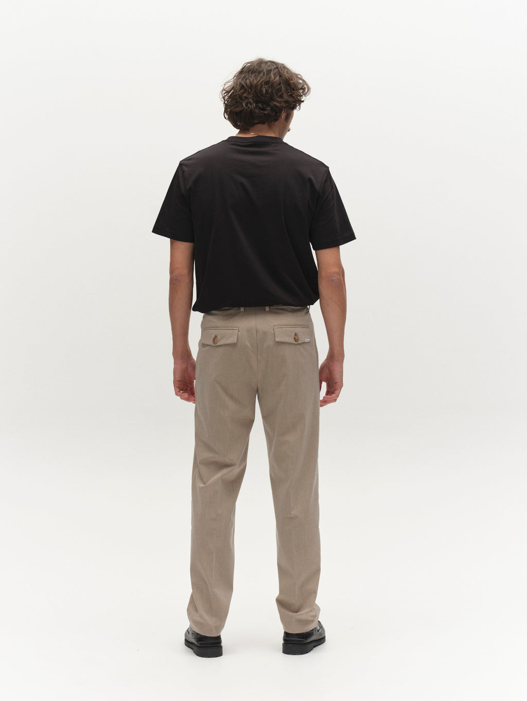 A person with short curly hair stands facing away, wearing a black short-sleeve shirt, Gabba GABjoe Tailor Dressy Pant in Sand with buttoned back pockets, and black shoes against a plain white background.
