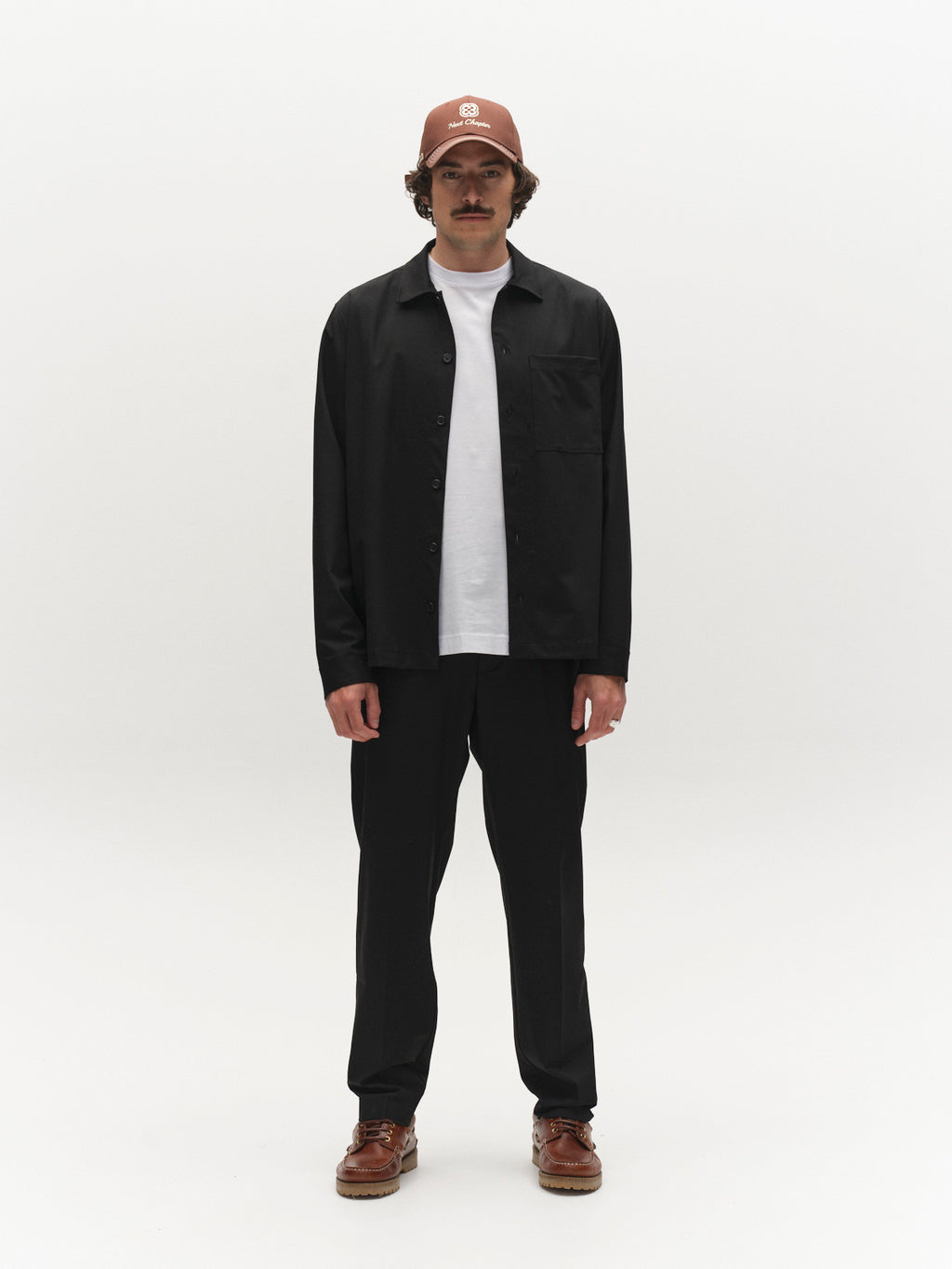 A man with a mustache faces forward wearing a brown cap, black button-up shirt over a white t-shirt, GABjoe Tailor Dressy Pant in Black by Gabba, and brown shoes, set against a plain white background.