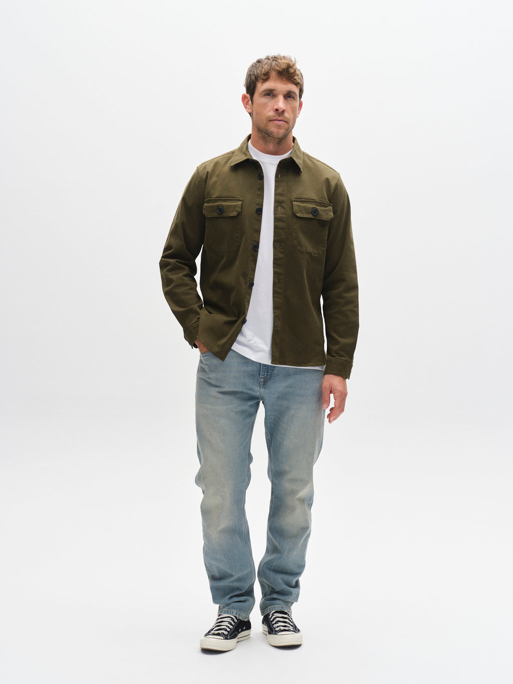 A man stands in a studio with a white background, wearing an olive green jacket over a white t-shirt, Gabba GABcarl CrosV2 Jeans in Lt. Blue Denim, and black sneakers.