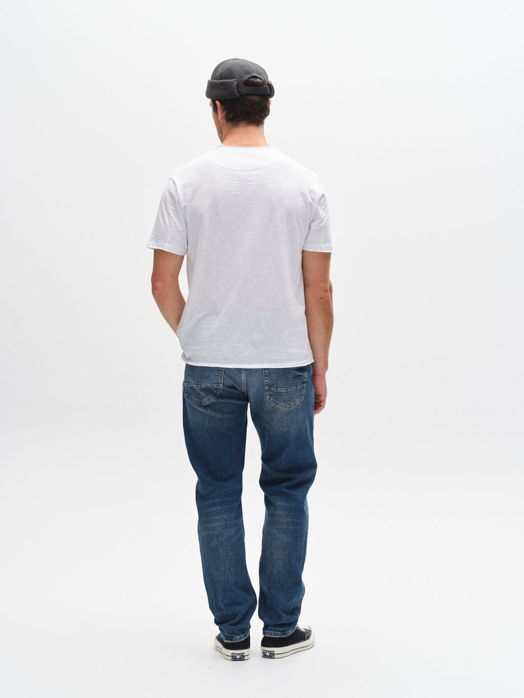 A man stands with his back to the camera, wearing a gray cap, white t-shirt, black sneakers, and Gabba GABcarl OskinV2 Jeans in dark blue denim against a plain white background.