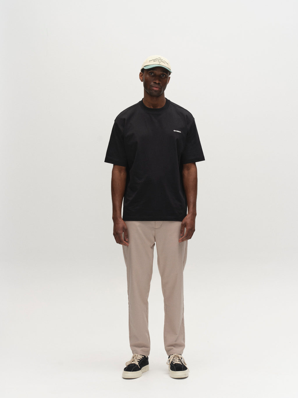 A man faces forward with a neutral expression against a white background, wearing the Gabba GABmonza Tasca Deme Pant in Birch, a beige cap, black short-sleeve t-shirt, and black sneakers with white soles.