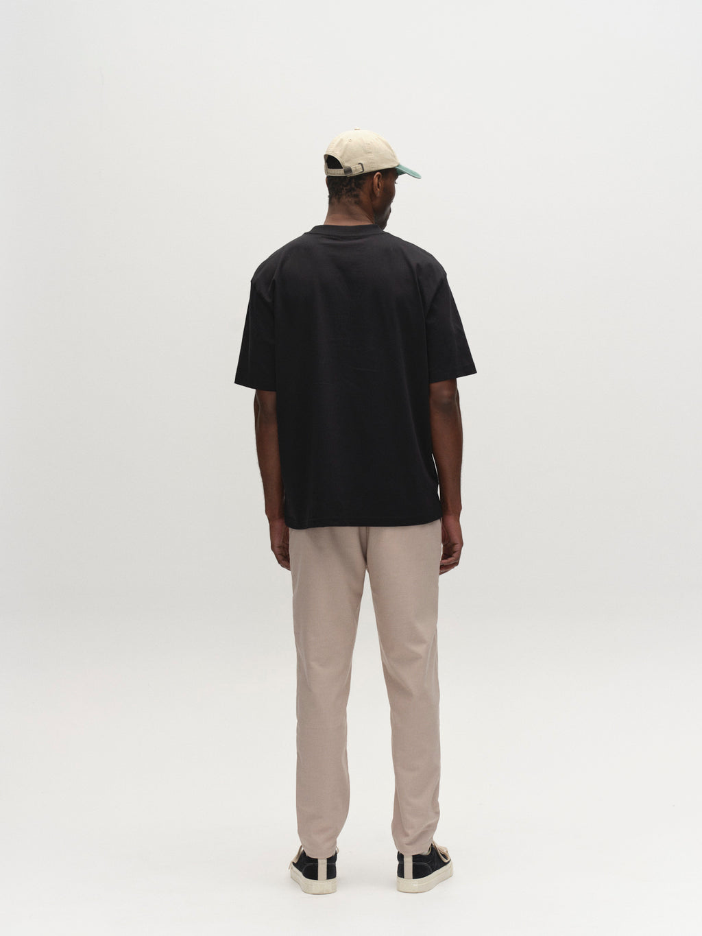 A man stands facing away from the camera against a plain white background, wearing Gabba's GABmonza Tasca Deme Pant in Birch, a black T-shirt, black and white sneakers, and a beige baseball cap.