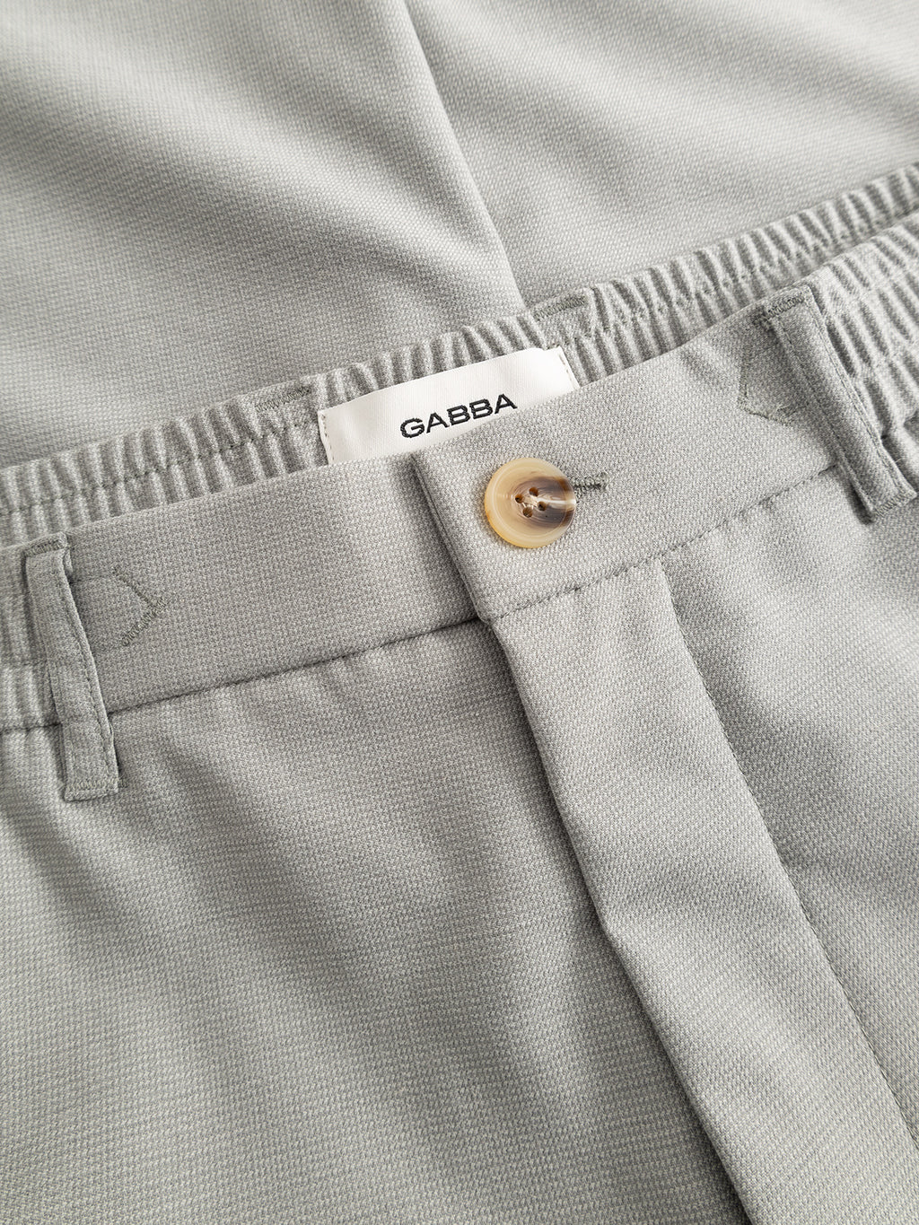 Close-up of the GABmonza Tasca Deme Pant in Mineral Gray by Gabba, featuring a beige button, elastic waistband, belt loops, and an inner white label with the brand name.