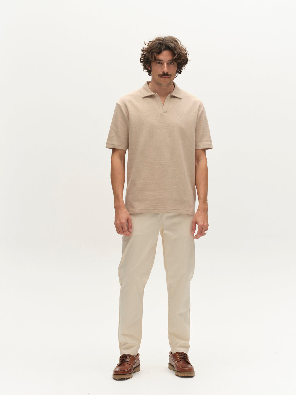 A man with curly hair and a mustache faces forward, wearing a beige short-sleeve polo, Gabba GABmonza Tasca Bolu TENCEL™ Pant - Birch with an elastic waistband, and brown shoes against a plain white background.