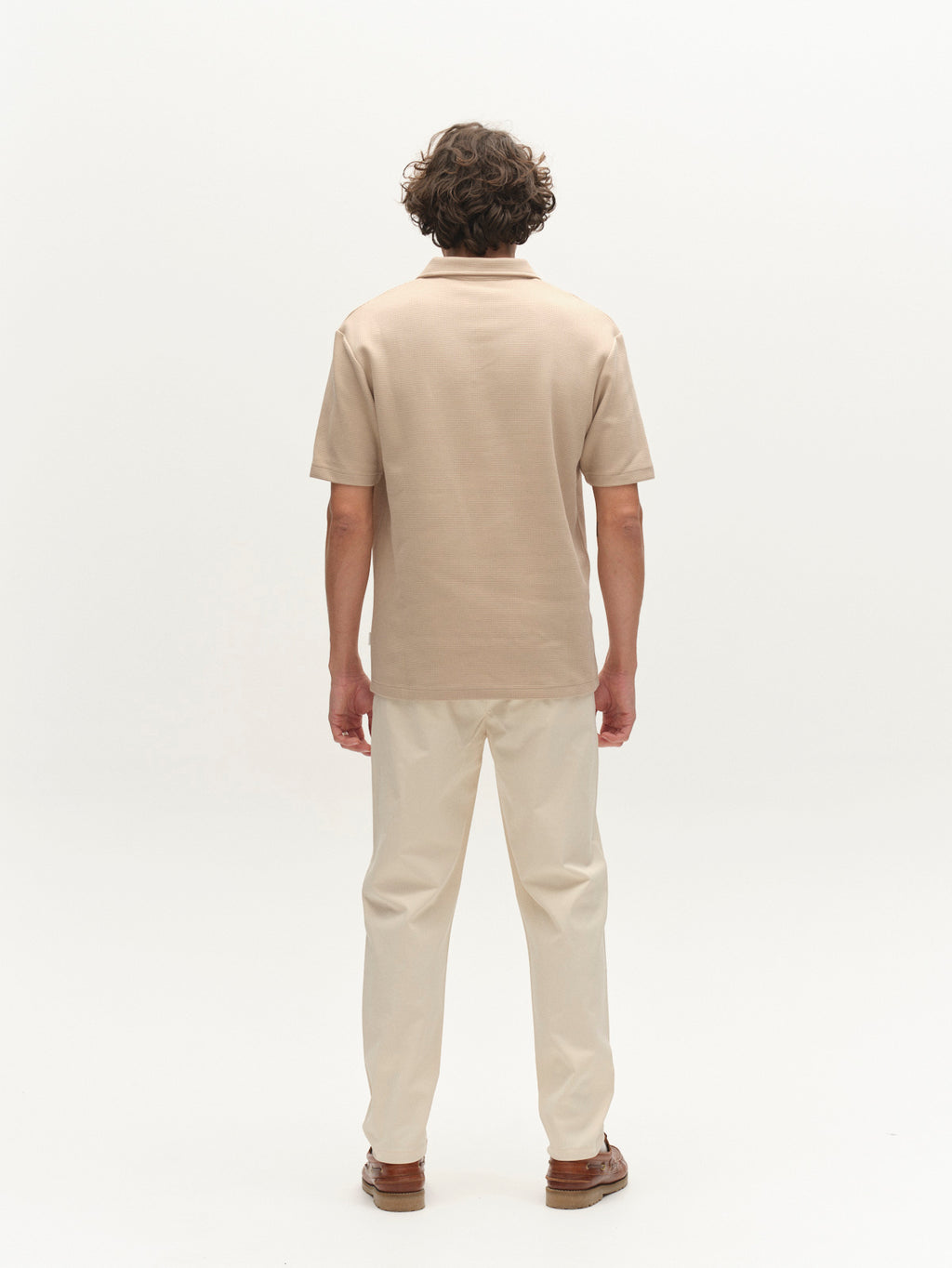A person with curly brown hair faces away, wearing a beige polo shirt, Gabba GABmonza Tasca Bolu TENCEL™ Pant in Birch (relaxed straight fit with elastic waistband), and brown shoes against a plain white background.